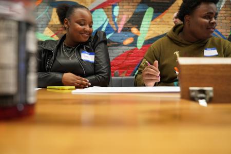 Two young adults, a woman named Mary and a man named Charles, sit side by side at a table, engaging in a group activity. Mary, wearing a black jacket and a 'Hello, my name is' sticker with her name written on it, smiles softly while looking down at the table. Charles, wearing an olive-green hoodie with a name tag, holds a pink pencil and appears focused on the task at hand. A vibrant mural featuring abstract shapes and colors is visible in the background.