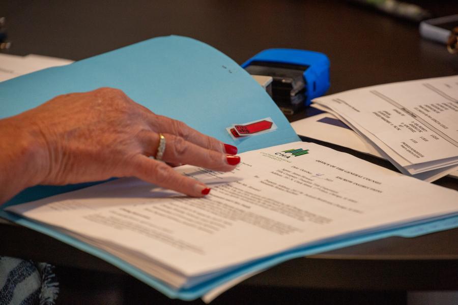 A close-up shot of a hand with red nail polish and a ring, organizing documents on a desk. The documents include official papers from the Chicago Housing Authority (CHA) within a blue folder. Office supplies, such as a stapler, are visible in the background.